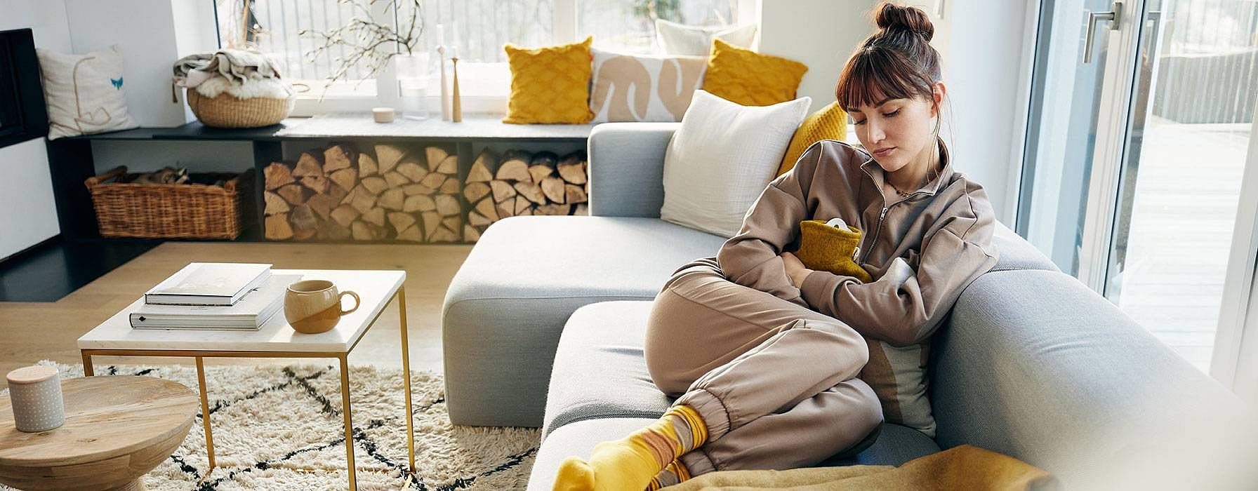 Frau mit Waermflasche auf dem Bauch auf einem Sofa