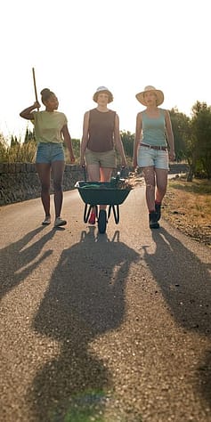 3 Frauen im Garten und eine schiebt einen Karren