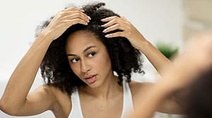 A woman with natural curly black hair looks at her reflection and checks her scalp, highlighting self-care and hair health.