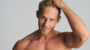 Laechelnder Junger blonder Mann mit Dreitagebart und unbekleidetem Oberkoerper, der sich mit der Hand durchs Haar faehrt
