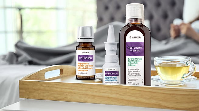 Hustensaft, Infludoron, Rhinodoron und Fiebermesser auf dem Tisch, Kind schläft im Hintergrund
