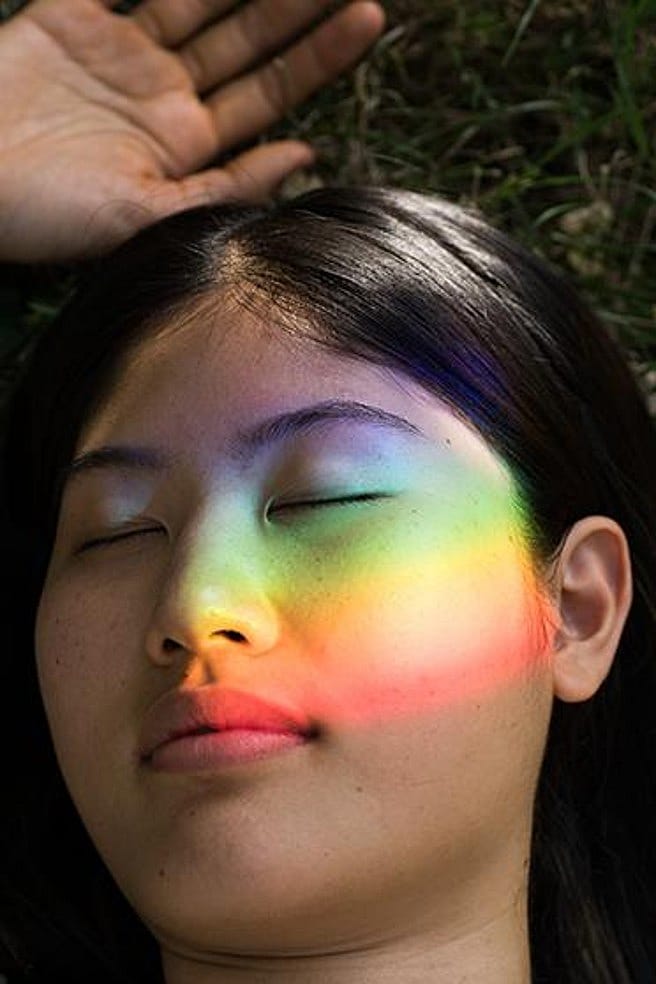 Frau mit geschlossenen Augen, im Gesicht die Spiegelung eines Regenbogenprismas
