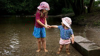 Bambino e bambina più grande con cappellini e piedi nel laghetto si scambiano qualcosa tra le mani