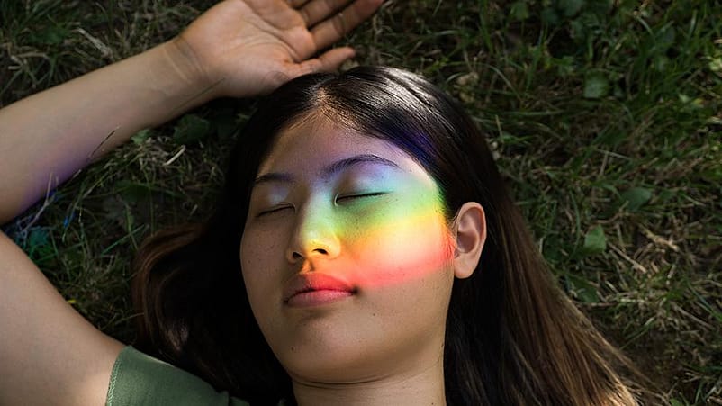 Ragazza sdraiata a terra con gli occhi chiusi e il riflesso dell'arcobaleno sul volto