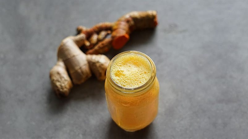 Glas mit Ingwer Shot neben einer Ingwerknolle und einer Kurkumaknolle