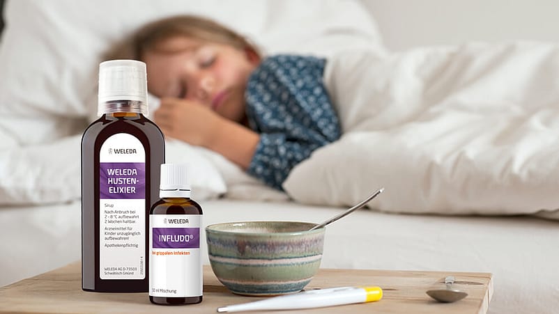 Hustenelixier, Infludo und Fiebermesser auf dem Tisch, Kind schläft im Hintergrund