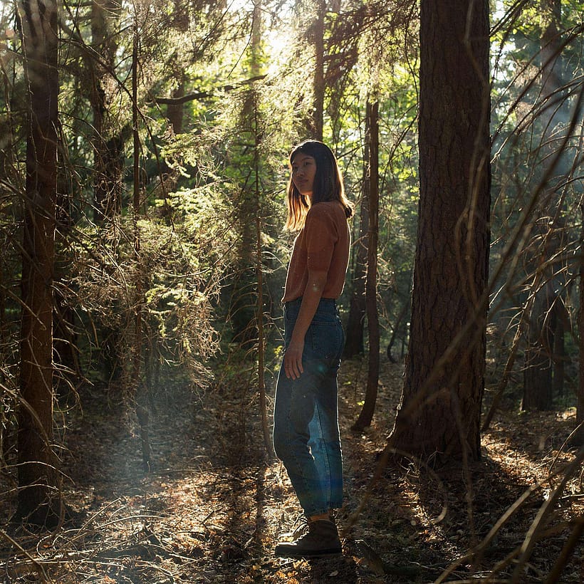 Frau steht in einem Wald, die Sonne strahle von hinten durch die Äste.