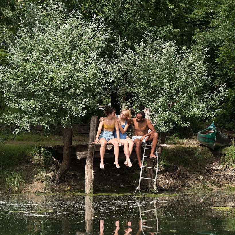 Zwei Frauen und ein Mann sitzen auf einem Badesteg am Wasser und umarmen sich