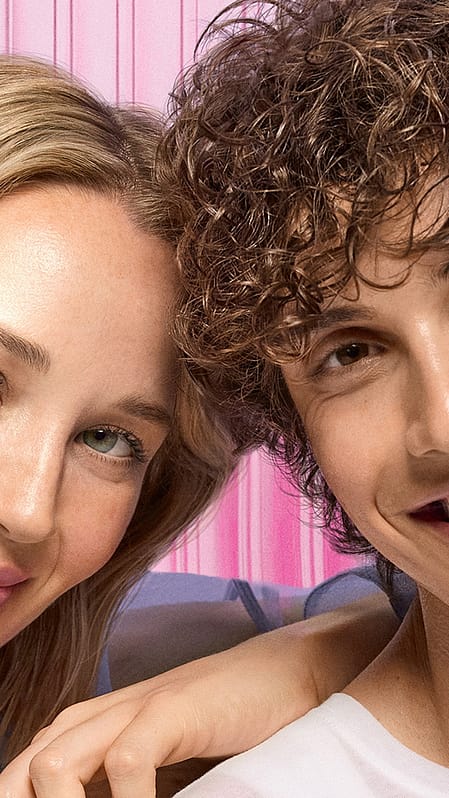 close-up of two smiling individuals with radiant, healthy skin, standing against a soft pink background. The woman has blonde hair and wears a sheer blue top, while the man has curly brown hair and wears a white shirt.