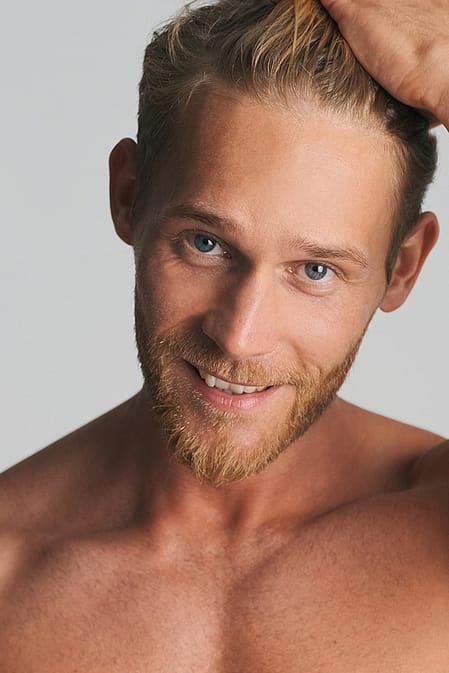 Laechelnder Junger blonder Mann mit Dreitagebart und unbekleidetem Oberkoerper, der sich mit der Hand durchs Haar faehrt