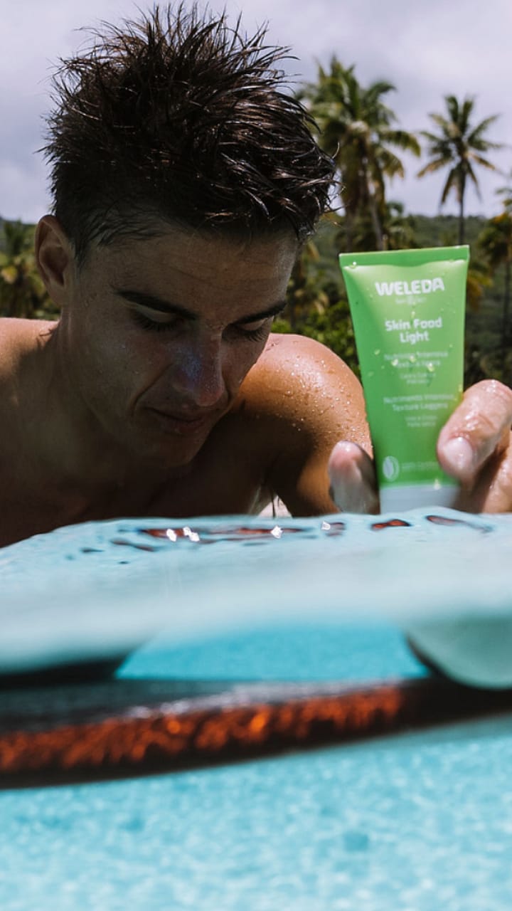 Hombre sobre una tabla de surf en el mar y sosteniendo una crema Skin Food de Weleda con la mano