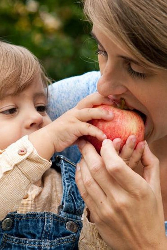 Mujer mordiendo una manzana que le da un niño pequeño que tiene cogido en brazos