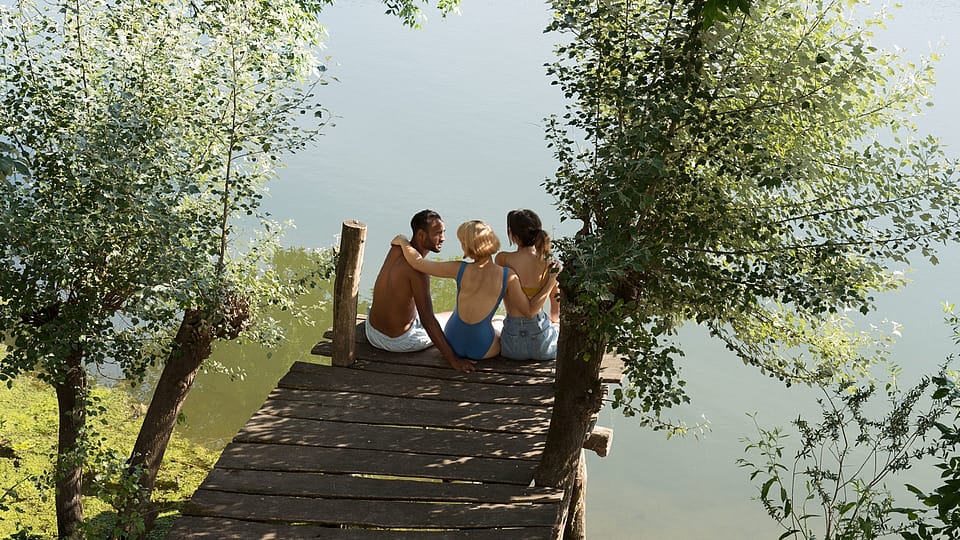 Zwei Frauen und ein Mann sitzen auf einem Badesteg am Wasser