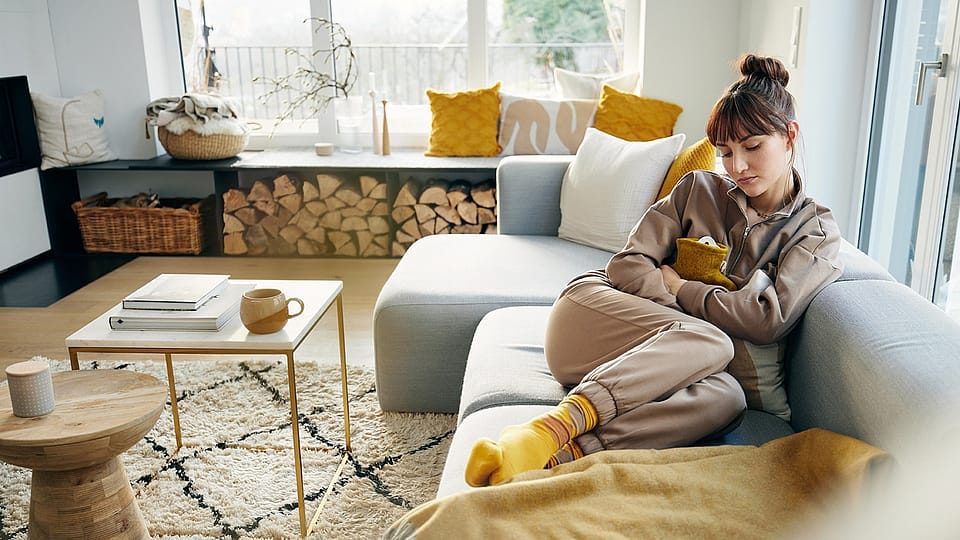 Frau mit Waermflasche auf dem Bauch auf einem Sofa