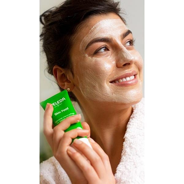 Woman holding a Skin Food tube with cream on her face as a skincare mask.