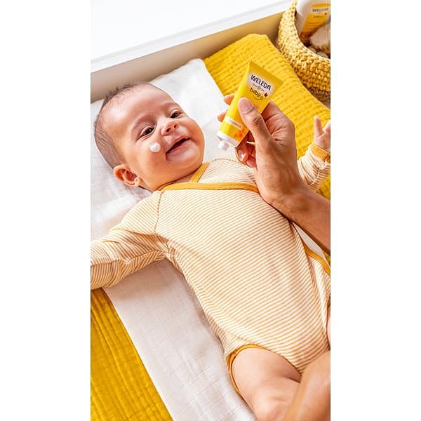 Smiling baby wearng yellow, lying down and being applied Calendula Face Cream on his face.