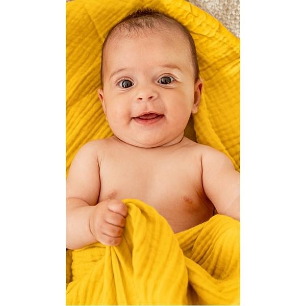 Smiling baby wrapped and lying down on a yellow muslin cloth.
