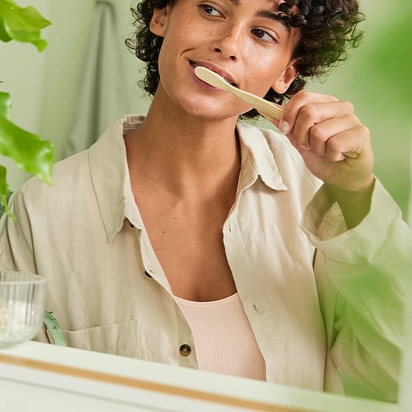 Dark skin woman with short curly hair brushing her teeth infront of the mirror.