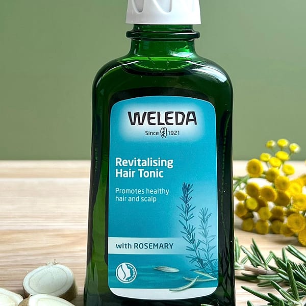 Revitalising Hair Tonic bottle on a sage green background with its ingredinets rosemary leafs and horseradish root.