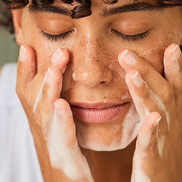 Dark skin woman with short curly hair with her eyes closed washing her face with a face cleasing foam.
