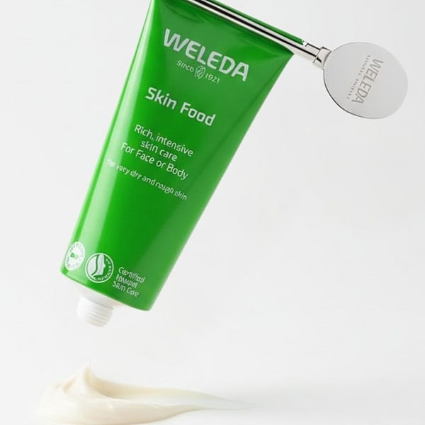 Weleda Skin Food tube with metal squeezer key, rich intensive moisturizer for face and body, certified natural skin care, cream swatch on white background.