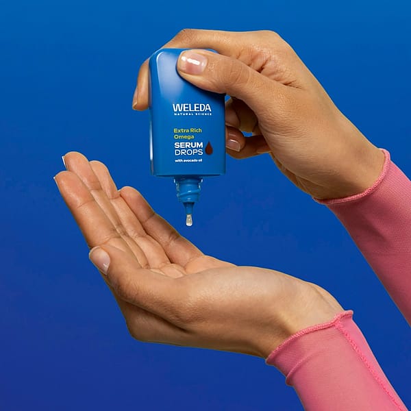 Hand applying Weleda Extra Rich Omega Serum Drops with avocado oil onto palm against a blue background – skincare product application for deep hydration and nourishment.
