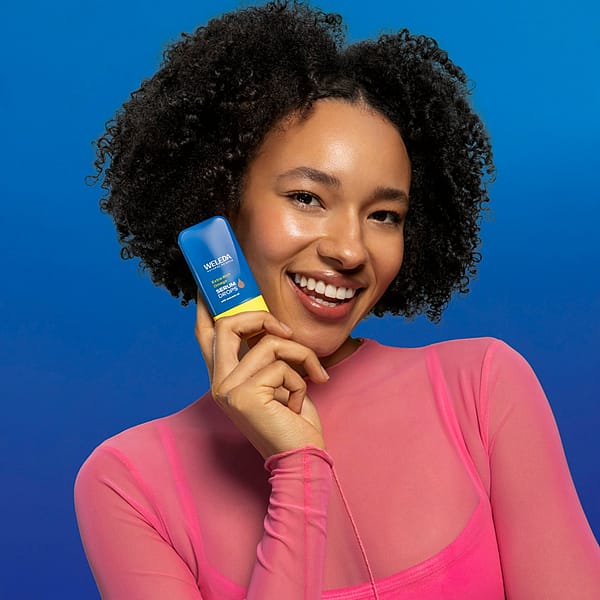 Smiling woman holding Weleda Extra Rich Omega Serum Booster Drops against a blue background, showcasing skincare product for deep hydration and radiant, healthy skin.